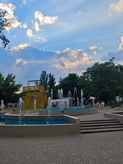 This is a photograph taken at the fountain complex in Taras Shevchenko Park, located in the city of Kryvyi Rih, Ukraine. The scene captures a vibrant public space during daytime, with a clear blue sky adorned with dramatic clouds. The foreground is dominated by a multi-level fountain with bright blue water, and water jets spraying upwards. Paved stone walkways form the ground surface. In the mid-ground, a person is partially visible standing near the fountain. Further back, a distinctive yellow wooden structure with multiple levels and platforms stands prominently, possibly a play area or observation deck, with a ring visible at the top. Several trees, including tall, slender ones and broader, leafy ones, line the background, creating a park-like atmosphere. A few people are seated on a bench in the distance, near some stalls or small structures, suggesting a recreational area. The overall impression is one of a lively and well-maintained urban park.