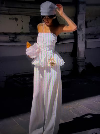 In this image, a person is lying down wearing a white dress and a white head covering. Their left arm is bent with their hand resting on their head, and their right arm is extended. A small, ornate purse is visible on the person's chest, attached by a thin chain. The background appears to be a dimly lit, rustic setting with crumbling stone or concrete structures and a dark, shadowed area. The lighting suggests an indoor or shaded outdoor environment, possibly at night or in low light conditions. The overall mood is somewhat somber or artistic, with the stark white of the clothing contrasting against the dark, weathered background. There is no visible text in the image. The image is oriented with the person lying down.