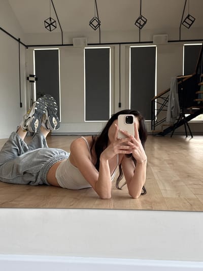 A young woman with dark hair is captured in a reflection, posing on a wooden floor in what appears to be a studio or home gym. She is wearing a white crop top and grey sweatpants, with athletic shoes on her feet. The woman is holding a smartphone and taking a selfie, with her face partially obscured by the device. Her pose is flexible and athletic, with her body contorted in a way that suggests she is engaging in a yoga or flexibility exercise.
The background visible in the reflection shows a clean, modern space with large windows or screens, some with black blinds. There are also geometric light fixtures hanging from the ceiling and a white speaker mounted on the wall. The overall setting suggests a well-lit, contemporary environment. The image is taken from a low angle, emphasizing the woman's pose and the reflective surface. No specific time of day or weather conditions are discernible. The woman's expression is not fully visible, but her posture conveys a sense of focus and athleticism. There is no visible text in the image.