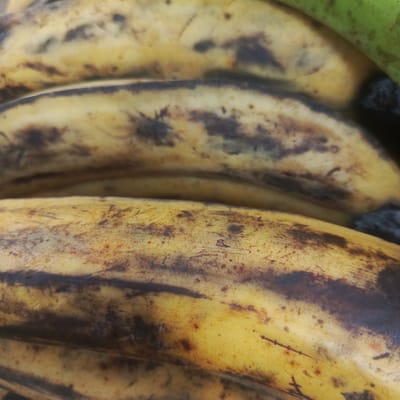 The image is a close-up, shallow-depth-of-field photograph of a bunch of ripe plantains. The plantains are yellow with brown speckling and some areas of darker brown bruising, indicating ripeness. The focus is primarily on the middle and foreground plantains, with the background slightly blurred. No specific location in Los Teques, Venezuela, is visible or suggested by the image content. There are no people, actions, or interactions depicted. The environment is simply the surface of the plantains.