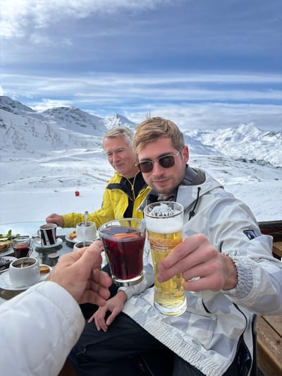 An outdoor scene in Saint-Martin-de-Belleville, France, depicts three individuals at an outdoor table on a snow-covered mountain with a vast, snowy mountain range under a partly cloudy blue sky in the background. In the immediate foreground, a person's left arm, clad in a white jacket, extends into the frame, holding a glass of dark red liquid containing orange slices, appearing to make a toast. To the right, a young adult male with blonde hair, wearing rectangular sunglasses with dark lenses and a grey and white camouflage-patterned ski jacket, holds a tall glass of light-colored beer, branded "BRASSERIE DU MONT BLANC," clinking it with the red drink. He is looking directly at the viewer with a slight smile. Seated to his left and slightly behind, an older adult male with short grey hair and a bright yellow ski jacket is visible, looking towards the left with a subtle smile. The clear glass tabletop features multiple white mugs, some with dark patterns, containing dark liquids and spoons, along with small plates holding pastries or snacks. A red ski lift chair and pole are visible in the mid-distance on the snow-covered slope to the left. The table is situated on a wooden deck or patio area.