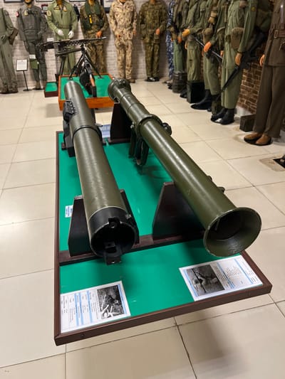 A museum exhibit features two green-painted rocket launchers mounted on a green background within a brown wooden frame. The launchers are displayed with information cards next to them. Mannequins dressed in various military uniforms stand to the left of the exhibit. The floor is light-colored tile.