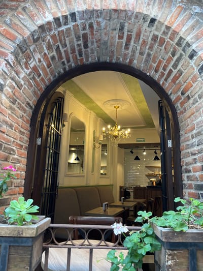 The image shows an arched brick entryway to a building in what could be Domingo Arenas, Mexico, although it is impossible to confirm that from the image alone. The archway is made of red and some darkened bricks and has a dark wooden frame. Through the archway, the building's interior is visible, featuring a chandelier and interior décor with green and white color schemes. Furniture, including tables and chairs, suggests the space is likely a restaurant. The view through the arch is oriented to make the viewer feel disoriented. A planter with succulents and flowers is visible on the right side of the archway.
