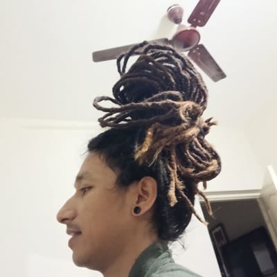 A side-view medium shot captures a person with dark hair styled in dreadlocks tied on top of their head. Some dreadlocks appear lighter brown. The person's face is visible in profile, featuring a noticeable nose and a slight smile. They have a black circular earring. The person is wearing a muted green shirt.

The background consists of a white ceiling with a brown ceiling fan, and a doorway leading to a dimly lit interior room. There is no information available to definitively place this photo in Bengaluru, India, but the photo was taken in India.