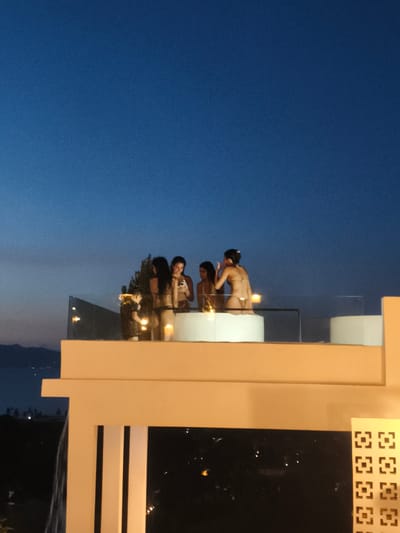A group of four women are gathered on a rooftop patio at dusk, standing around a white, circular hot tub. The sky is a deep, clear blue, transitioning to a lighter hue near the horizon where mountains are visible in the distance. The women are wearing bikinis and appear to be engaged in conversation and leisure. One woman in the foreground is looking at her phone, while another stands next to her, looking in the same direction. The patio has a glass railing overlooking a city with lights visible in the darkening landscape. A patterned decorative panel is visible on the right side of the image, suggesting a modern architectural style. The scene is illuminated by ambient light from the sky and some artificial lighting, including candles placed on the edge of the hot tub. The location is identified as Baan Bang Rak, Thailand.