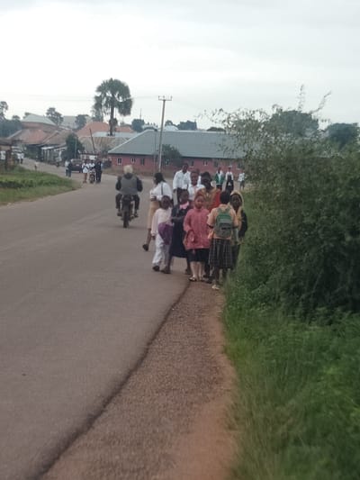 The image captures a group of people, including children, walking along a paved road in what appears to be a rural or semi-urban setting in Du, Nigeria. Several children, some wearing school uniforms and carrying backpacks, are prominent in the foreground and mid-ground. A person is riding a motorcycle in the mid-ground, and other individuals are visible further back, walking or standing.

The scene includes buildings with red brick walls and tiled roofs in the background, suggesting a village or small town. A tall palm tree is also visible, a common sight in many parts of Nigeria. The weather appears to be overcast, indicated by the bright, diffused light and the muted colors of the sky. The overall impression is one of daily life, possibly the start or end of a school day, with people going about their business. There is no visible text in the image.