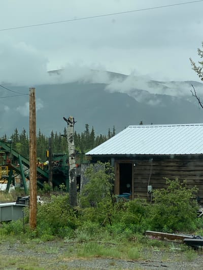 The image shows a rustic wooden building with a corrugated metal roof, situated in a rural or semi-industrial environment. In front of the building, there is dense green foliage and a dead, white-barked tree. To the left of the building, a section of industrial machinery, likely for mining or processing, is visible, painted in green and yellow. A wooden utility pole stands in the foreground, to the left of the dead tree. In the background, a range of mountains is shrouded in low-lying clouds, with a dark, overcast sky above. The scene appears to be in Haines Junction, Canada, given the characteristic landscape of mountains, boreal forests, and the presence of infrastructure related to resource extraction. There are no people visible in the image.