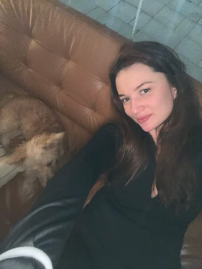 A woman with long dark hair lies on a tufted brown leather couch, looking at the camera. She wears a long-sleeved black top. A small, furry dog curls up near her on the couch, blending into the background. The picture is taken at an angle, with a hint of an off-white surface in the background. The lighting is somewhat muted.