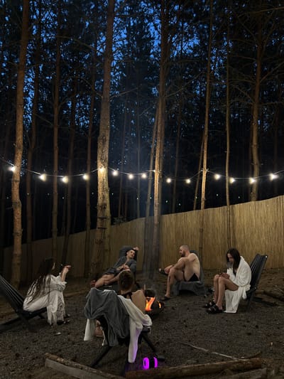 In Ivachiv Horishnii, Ukraine, at night, a group of five individuals are gathered around a campfire in a forested area. String lights are suspended between trees, illuminating the scene. Two individuals are seated in chairs, facing the fire, while one person lies on the ground. Another person is seated in a chair, looking at their phone, and a fifth person is seated in a chair, appearing to smoke. The ground is covered in gravel, and a bamboo fence is visible in the background.