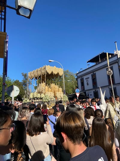 The image captures a religious procession in Córdoba, Spain, likely during Holy Week (Semana Santa). A large crowd gathers along the street, primarily composed of adults. At the center is a richly adorned float or "paso," embellished with gold accents, flowers, and candles. The float may carry a religious icon or statue, but that is not visible. Participants in the procession surround the float, some wearing traditional robes and conical hats. A sign with two arrows is visible on top of the crowd. Street signs are visible on a building behind the crowd and the float, reading "Meson El 6" and "Puerta Nueva." The environment includes buildings, street lamps, traffic signals, and a clear, blue sky.