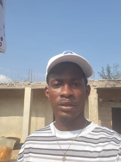 In Egbagu, Nigeria, a man is captured in a selfie, his face partially obscured by a white baseball cap with a blue logo. He is wearing a horizontally striped t-shirt in shades of gray and white, with a thin gold chain and a cross pendant visible. The background reveals an unfinished building with a concrete ledge adorned with exposed rebar and thorny branches, under a clear blue sky. To the left, a clear plastic cup filled with orange-colored food items sits next to a woven basket.