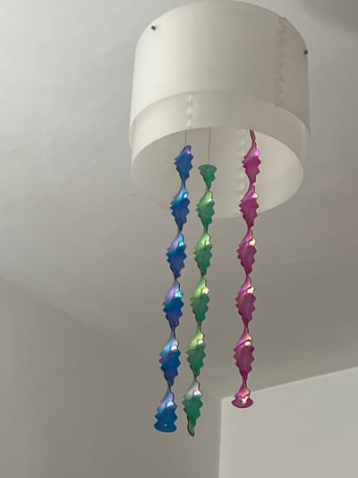 A white cylindrical object is mounted on a white wall, with three iridescent, decorative strands extending horizontally from it. The top strand is pink, the middle is green, and the bottom is blue. The location is Picassent, Spain.