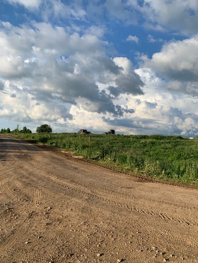 The image depicts a dirt road curving through a rural landscape under a dramatic sky filled with dark, heavy clouds interspersed with patches of blue. The road is textured with tire tracks and scattered pebbles. To the right of the road, a field of lush green grass and vegetation stretches towards a distant horizon. In the middle ground, several dark, silhouetted structures, possibly farm buildings or hay bales, are visible on the elevated terrain. A large, solitary tree stands prominently in the grassy field. The overall scene suggests a natural, undeveloped environment in the vicinity of Святица, Russia, possibly in the summer or early autumn given the green foliage and cloudy sky.
