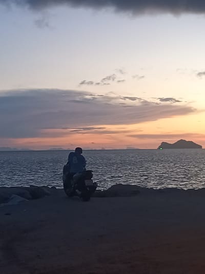 The image depicts a serene sunset scene over the sea. In the foreground, a person is seated on a motorcycle on a beach. The person is silhouetted against the dusky sky, wearing a dark top with what appears to be a number "8" on the back. The motorcycle is also dark and facing away from the viewer. The beach itself is rocky and sandy. The sea is calm with gentle waves, and in the distance, several islands are visible on the horizon. The sky displays a gradient of colors, from pale blue at the top to warm oranges and pinks near the horizon, with scattered clouds adding texture. A small, bright green light is visible near one of the distant islands. The overall atmosphere is tranquil and reflective. The scene is consistent with a location in the Ban Tai Subdistrict of Thailand, known for its beautiful beaches and sunset views.