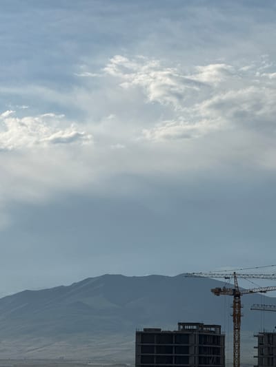 The image depicts a construction site in Abovyan, Armenia, with mountains in the background. In the foreground, there are two partially constructed buildings, one of which is a concrete frame structure with multiple floors. Yellow construction cranes are present, with one prominent crane positioned between the buildings. The sky is partially cloudy, with soft white clouds against a pale blue backdrop. A small, dark bird is visible in the sky, adding a touch of life to the scene. The overall atmosphere is one of urban development against a natural mountainous landscape.