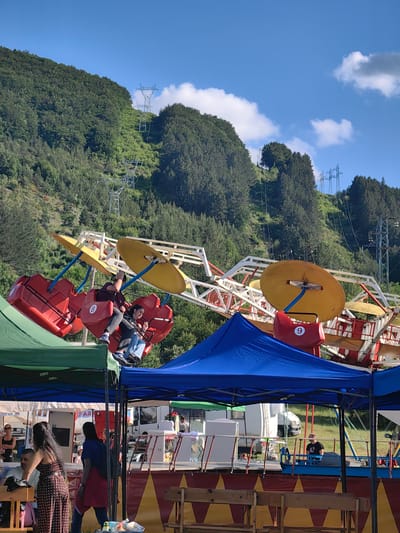 The image depicts an outdoor amusement park scene, likely during daytime given the bright sunlight and blue sky with scattered clouds. In the foreground, the tops of two tents, one green and one blue, are visible. Behind these tents, a large amusement park ride is in motion. This ride features red gondolas suspended from a yellow and white mechanical structure. Two people are visible in one of the gondolas, seemingly enjoying the ride. The background of the image is dominated by a lush green, tree-covered hillside. High tension power lines and their supporting pylons are prominently featured traversing the hillside, indicating the location's proximity to a power infrastructure. The overall impression is of a recreational area situated in a natural, possibly mountainous, environment with evidence of significant electrical infrastructure. The location is identified as the city of Lozen, Bulgaria.