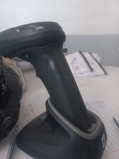 The image shows a close-up, angled view of a dark grey barcode scanner docked in its charging base. The scanner has a curved handle and a wider top section with a visible scanning window and indicator lights. The base is also dark grey with a lighter grey trim around the docking area. To the left of the scanner, a black, crinkled object, possibly a plastic bag, is visible, along with the tip of a silver pen. In the background, out of focus, are stacks of white papers with black markings and what appears to be a white wall. The scene suggests an indoor setting, likely an office or retail environment. The metadata does not contain information about the location in the city of Yorkatay, Uzbekistan, as this is an image analysis task.