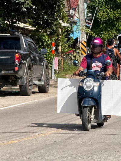A man wearing a pink helmet and a t-shirt with a crown design is riding a dark blue Honda scooter on a paved road in Ban Tai Subdistrict, Thailand. He is carrying a large white panel horizontally in front of him. To the left of the scooter, a black pickup truck is parked on the side of the road. In the background, there are trees, buildings, and a utility pole with wires. The sun is shining, casting shadows on the road. Another person on a scooter is visible in the background to the right of the main subject.