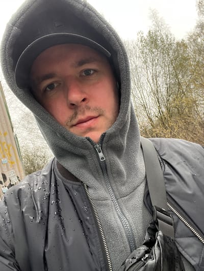 The image is a rotated selfie, oriented vertically and slightly counter-clockwise, showing a young man outdoors. He looks directly at the camera with a neutral expression. He is wearing a grey hooded fleece jacket, with a "YKK" logo visible on its zipper pull, layered under a black puffer-style jacket. A black baseball cap is worn beneath his hood. Both his black jacket and grey hood are covered in numerous water droplets, suggesting it is raining or has recently rained. A black strap from a bag, possibly a fanny pack, is visible over his shoulder, with a plastic buckle, and a portion of the black, shiny, and wet bag is seen below it.

The background features an overcast, light-colored sky and a dense canopy of trees with mostly bare branches, though some faint light green foliage is visible, indicating early spring. Below the trees, at the very bottom of the frame, there is a hint of a structure or wall with graffiti. Text visible on this structure includes "ATOME" and "O 7074". The scene depicts an outdoor setting in Odintsovo, Russia, under diffused, cloudy lighting conditions.