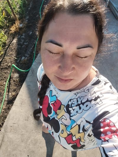 A woman with dark hair and closed eyes is pictured from a high angle, looking down. She is wearing a white t-shirt with a colorful Mickey Mouse pattern and the word "Disney" in black script. The woman has fair skin with freckles. Her dark eyebrows are well-defined. Her hair is pulled back into a braid that falls over her right shoulder. She is outdoors on a sunny day, standing on a concrete surface adjacent to a patch of dirt and dry grass. A green hose snakes across the dirt area. The background is out of focus, suggesting a rural or suburban setting, possibly in Krasny Gulyai, Russia.