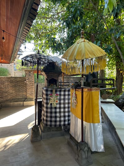 The image shows an outdoor scene in Kediri, Indonesia, with two ceremonial umbrellas standing in front of a small altar-like structure. The altar is draped in black and white checkered fabric, with a smaller yellow and white draped structure to its right. Both umbrellas are ornately decorated with fringe. The environment suggests a temple or a place of worship, with lush greenery visible in the background. The ground is paved with light-colored tiles, and a brick wall is visible to the left. The lighting indicates it is daytime, with sunlight casting shadows on the ground.