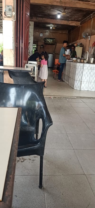 This is a vertical, eye-level, medium shot taken outdoors in front of a food stall. The foreground is dominated by the backs of two black plastic chairs and a table. The chairs are worn and have visible scratches. In the mid-ground, a young girl in a pink and white patterned dress is standing at the counter of the food stall, looking down at something in her hands. She has dark hair pulled back in a ponytail. To her left, a person wearing a black headscarf and black clothing is leaning over the counter, also seemingly engaged with the items on the counter. To the right of the girl, a young man in a teal t-shirt and dark pants stands behind the counter, looking towards a man in a dark green t-shirt who is also behind the counter, appearing to be serving a customer or preparing food. The food stall counter is tiled with a gray and white patterned mosaic. Behind the counter, various kitchen appliances and equipment are visible, including pots, pans, and what appears to be a refrigerator. A television screen mounted on the wall shows a nature scene. The environment appears to be casual and busy, typical of a local food stall. The floor is tiled with light gray squares. The ceiling is made of rough, unpainted material and has fans. The overall impression is a slice of everyday life at an Indonesian food establishment.
