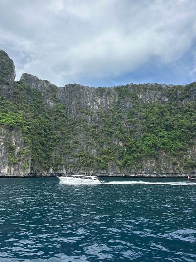 A speed boat moves through dark blue water in front of a sheer cliff face covered in green foliage. The cliff face is rocky and uneven, with many indentations and ledges. The sky above is cloudy, with patches of blue sky visible. The boat is white with a red stripe and has several passengers visible inside. The water is choppy, with white wakes trailing behind the boat. The scene is serene and picturesque, suggesting a tropical location. The location is Haad Hin Khom, Thailand, known for its dramatic limestone cliffs and clear blue waters.