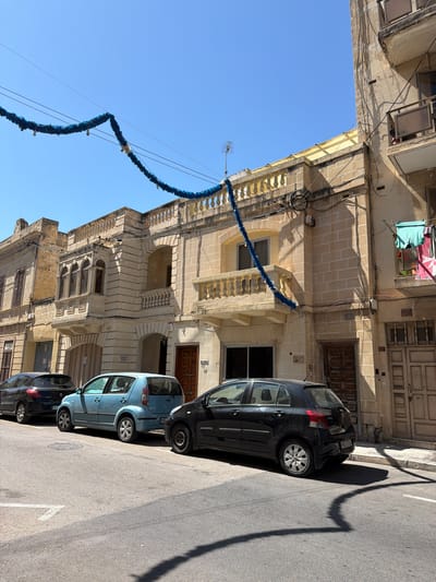 In Pietà, Malta, along a sun-drenched street, a row of three cars is parked on the right side of the road. The vehicles, a black Peugeot, a light blue hatchback, and a black Toyota Yaris, are aligned on the asphalt. Adjacent to the parked cars, a row of stone buildings stands, showcasing architectural details typical of the region. Blue garlands are strung across the street and draped over the buildings, possibly for a festive occasion. The buildings feature balconies, balconies with intricate balustrades, and windows with shutters. Laundry hangs from a balcony on the rightmost building. The sky above is a clear, vibrant blue.