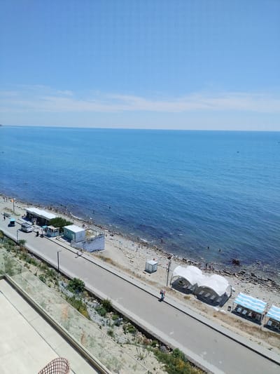 This is an aerial, wide-angle, full shot of a sunny day at a beach resort in Anapa, Russia. The scene captures the vast expanse of the blue Black Sea meeting a light blue sky. The shoreline is a mix of sand and pebbles, with a road running parallel to it.
Along the beach, several white, tent-like structures are set up, likely for shade. Some beach chairs are visible beneath these structures. There are a few people scattered on the beach, some swimming in the sea and others sunbathing. A small boat is visible in the distance on the water.
The road has a few cars and motorcycles parked along its side, as well as some small buildings that appear to be kiosks or changing rooms. A few individuals are walking or cycling along the road and the beach path. The foreground shows a modern balcony with a glass railing, overlooking the beach and sea, suggesting the vantage point is from a hotel or residential building. The vegetation is sparse, with some dry grasses and low-lying plants along the embankment. The overall atmosphere is one of a typical summer day at a seaside resort.