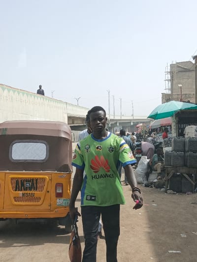 The image portrays an outdoor street scene in Fagge C, Nigeria, under a bright, hazy sky. A male subject, appearing to be in his 20s, with dark skin and short black hair, is positioned centrally, walking towards the viewer. He wears a short-sleeved jersey that is predominantly light green with blue accents, featuring "HUAWEI" and a red floral logo on the chest, alongside "NRL" and "ISC" branding. He wears dark trousers and a dark bracelet on his right wrist. He carries a brown bag in his left hand and a small pink object in his right hand.
To the subject's left, a yellow and brown motorized tricycle (keke napep) is parked, facing away from the camera. Its rear features the text "HISABI," the license plate "FGE-43JN," and markings "AG 01M" and "RE." Behind the tricycle, a light-colored concrete wall, approximately 1.5 meters high, is visible with a green decorative pattern along its top edge. A distant male figure in dark clothing stands atop this wall.
To the subject's right, the scene transitions into an unpaved market area. A teal market umbrella shades a stall. Multiple black plastic jerrycans, each marked with "25," are stacked on a wooden stand. Several individuals are present in the background, some near white sacks on the ground. Partial construction scaffolding is visible on a building in the far right background. Elevated road infrastructure with lampposts runs horizontally across the mid-background. The ground consists of dirt or sand.