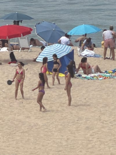 The image depicts a crowded beach scene on a sandy shore with the ocean in the background. Several people are visible in swimwear, engaged in various activities. In the foreground, a woman in a pink bikini stands holding a black object, possibly a small ball or paddle. To her right, another woman in a purple bikini is visible. Further back, a woman in a pink bikini stands with her back to the camera, and to her right, a woman in a black bikini is also visible. Scattered throughout the beach are several beach umbrellas in red and blue. Some individuals are lying on towels or chairs, while others are standing or walking. The overall atmosphere suggests a sunny day at the beach with many visitors. The location is identified as the city of Espinho, Portugal.