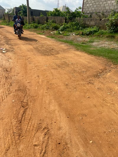 An adult male is riding a motorcycle on a dirt road in Dam Camp, Nigeria. He is wearing a dark blue shirt and pants. A young child, wearing a white shirt and a red bowtie, is seated in front of him on the motorcycle. They are both looking forward. The road is made of orange dirt and has tire tracks. There is green grass and vegetation on the right side of the road. In the background, there are buildings and a wall. The sky is cloudy.
