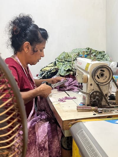 This media file shows a woman in Habaraduwa, Sri Lanka, working at a sewing machine. She has her hair pulled up in a bun and is wearing a maroon top. A measuring tape is wrapped around her neck. She is holding a pair of scissors in her right hand and appears to be cutting fabric. The sewing machine is a white Singer model, with several spools of thread and fabric scraps visible around it. The background is a simple white wall, and the scene is indoors. The lighting suggests it could be daytime. The overall impression is one of focused work in a home or small workshop setting.