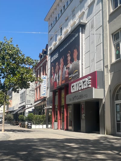 The image captures a street scene, notably featuring the Camera arthouse cinema in Saarbrücken, Germany. The cinema's signage is prominent, along with an advertisement poster displaying a movie. A business sign reading "KRAEMER HERRENMODE" is also visible. Additional elements include other businesses, such as a Trattoria restaurant, trees, and sidewalk features like seating arrangements. The sky is bright blue, adding to the daylight setting of the scene. Shadows cast across the pavement further suggest a sunny day.