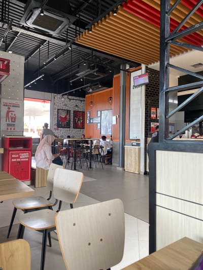 This image shows the interior of a fast-food restaurant, possibly KFC, in Lhoksukon, Indonesia. The scene is captured from a high angle, looking down into the dining area. Several people are visible, some sitting at tables and others standing. The decor features a combination of modern and rustic elements, with wooden slats on the walls and ceiling, black metal accents, and patterned wallpaper. A large digital menu board is mounted on the wall, displaying text in what appears to be Indonesian. Signs on red bins also have Indonesian text, promoting a "clean up yourself" culture.
The overall atmosphere appears to be casual and busy, suggesting it's a typical day at a popular eatery. There are rows of chairs and tables set up for customers. Natural light filters in from a large open doorway or window on the right side of the image, illuminating the dining area. The presence of the digital menu and promotional posters indicates that it's likely daytime, with customers coming and going. The restaurant seems to be a well-lit and inviting space.