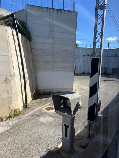The image depicts an outdoor scene, likely a railway or industrial area, viewed from what appears to be a vehicle. The setting is under bright, clear daylight with a blue sky and scattered white clouds.
In the foreground, a gray metal post features a black 'T' symbol on a white rectangular panel. Adjacent to it, a gray metal housing, possibly for a signal or part of a barrier mechanism, is visible. Stretching across the midground is a railroad crossing barrier arm, distinguished by its black and white diagonal stripes, which appears to be in a partially raised or open position. Above the barrier, a metallic lattice structure, possibly an overhead line support, stands against the sky.
The background is dominated by a series of tall, light gray concrete walls, some exhibiting patches of white paint or repairs. Green foliage and weeds grow at the base of these walls. A wire mesh fence is visible on the far left. The ground consists of dark asphalt pavement on the right and lighter concrete paving on the left, where a black pipe runs along the base of a wall. Faint white handwritten text, "2016 BIG," is visible on the concrete ground near the pipe. No activity or people are present, indicating a static moment in an infrastructure-heavy zone, consistent with a location like Bari, Italy.