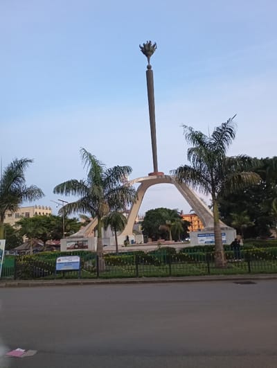 The image shows a tall, thin, concrete monument with a flame-shaped top located in a public square in Arusha, Tanzania at the coordinates -3.369, 36.688. The monument is constructed of concrete and has a tall, slender structure with a large, open arch at the bottom. The arch is constructed of concrete and has a light tan color. The top of the monument is decorated with a flame-shaped structure made of metal. The monument is surrounded by a black metal fence and lush green bushes.  A small, blue sign is visible on the right-hand side of the monument. In the background, a few buildings and palm trees can be seen. Two people are walking across the square in the distance. The sky is a clear blue, indicating the time of day is likely late afternoon.