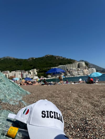 The image captures a sunny beach scene in Rafailovići, Montenegro. In the foreground, a white baseball cap, embroidered with "SICILIA" in black capital letters and featuring a circular Italian flag emblem, rests on prominent greenish-blue rocks. Adjacent to the cap is a clear bottle with a yellow label indicating "MANGO & LIME FLAVOURED REFRESHING CIDER." The ground consists of a pebbly beach with stones of varying hues. The midground shows numerous individuals relaxing under blue and light blue umbrellas on the blurred beach, with the calm sea visible beyond. The background features green, tree-covered mountains rising behind a dense array of multi-story buildings, likely hotels or residential structures, lining the coastline under a clear, cloudless blue sky.