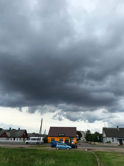 This is a full-frame, eye-level shot of a street scene in Porėčča, Belarus, under a heavily overcast sky. The foreground is dominated by green grass and a dirt path winding towards the right. In the mid-ground, a road runs horizontally, bordered by a sidewalk and several buildings.
On the left, a long, traditional red wooden house with white-trimmed windows and a grey roof is visible. Further along the road, a white minivan is parked. Adjacent to it is a vibrant orange, single-story commercial building with a brown roof, featuring a sign with Cyrillic text that reads "ГРЕНКА". In front of this building, a blue minivan and a dark blue sedan are parked.
To the right of the orange building, a paved road curves to the right, leading further into the settlement. A grey wooden house with a grey roof and a chimney stands on the right side of the road. A blue sign with a white pedestrian symbol is visible on a post near this house. The background consists of trees and more buildings, indicating a residential or mixed-use area. A dark, ominous cloud formation covers the upper half of the image, suggesting an impending storm.