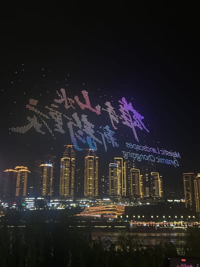 A nighttime drone light show is taking place above a cityscape featuring illuminated skyscrapers and a river. The drones are arranged to form large Chinese characters that appear to say "壮丽山河" (magnificent mountains and rivers) and "重庆" (Chongqing). Below the drone display, the buildings are lit up with yellow and white lights, and the river reflects the city lights. A portion of the text "Fantastic Chongqing" is also visible, spelled out in English by the drones, but it is inverted. The scene is located in the Jiangbeicheng Street (江北城街道) area of Chongqing, China.