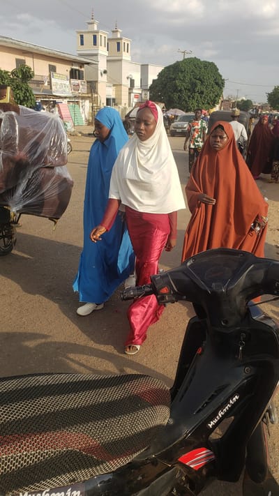 The photograph, taken in Kawo, Nigeria, shows three women dressed in traditional Muslim attire. The woman closest to the camera is wearing a blue hijab and is seated near a pushcart loaded with furniture wrapped in plastic. Adjacent to her is another woman in a white hijab and a red garment. Further back, the third woman is wearing a brown hijab. They stand on an asphalt road. To the right is a black motorcycle with the inscription "Mushmin" visible. In the background, buildings and trees are visible, including what appears to be a mosque with distinct towers. People are blurred in the distance.