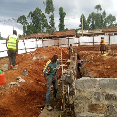 This image captures a vibrant construction scene in Borabu Chitago ward, Kenya, under an overcast sky. Several individuals are engaged in building activity on a site characterized by significant earthworks and reddish-brown soil.
In the foreground to the left, a man wearing a bright yellow safety vest with "FIRST GROUP SOLUTIONS LIMITED" printed on the back walks away from the camera, wearing light-colored shoes or barefoot. Near him, an orange container rests on the ground. To his right, another man in a green and yellow t-shirt, bearing a "Vodafone" logo, stands near a newly constructed stone and mortar foundation, looking downwards, seemingly focused on the work.
Further into the site, three men are actively working on building foundations. Two are positioned within a trench, meticulously laying stones and mortar, while another stands above, observing or assisting. Rebar is visible, indicating structural reinforcement, and string lines are used to ensure alignment.
On the right side of the frame, a woman in an orange top, brown skirt, and blue headscarf stands atop a mound of excavated earth, observing the construction. A yellow safety helmet rests on the ground near her. The ground is a rich, red-brown soil, with scattered stones and construction debris.
In the background, a temporary fence made of corrugated iron sheets and wooden posts delineates the construction area. Beyond the fence, simple, single-story buildings with corrugated iron roofs are visible. One building displays a sign with partially legible text, appearing to read "UTUMIS CYBER & SALO" (likely "SALON"). Tall, slender trees punctuate the skyline, contributing to the natural backdrop of this busy construction environment.