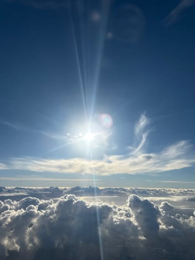 This image presents an aerial view of a bright, sunlit sky and dense cloud formations, purportedly located over Amora, Portugal. The scene is dominated by a clear blue sky in the upper half, with the sun prominently positioned in the upper-left quadrant. The sun emits strong, radiant light rays that traverse diagonally downwards, creating distinct lens flares, including a noticeable green hue, against the blue expanse.

Beneath the upper sky, a vast, undulating layer of white and grey cumulus clouds extends across the lower half of the frame, appearing to form a horizon line below the camera's viewpoint. These clouds are brightly illuminated from above by the sun, showcasing defined contours and deep shadows, indicating significant depth and volume. Some wispy, lighter clouds are visible at mid-level in the sky above the main cloud layer, particularly towards the right. No discernible landmass, structures, or individuals are visible beneath the cloud cover or in the immediate environment. The overall impression is one of high altitude and expansive open sky on a clear day.