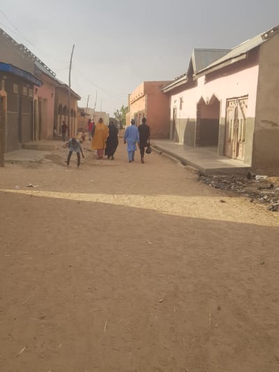 The image depicts a street scene in Dutsinma B, Nigeria. A group of people, dressed in various attires including blue and yellow robes, are walking down a dirt road lined with buildings made of light-colored materials. The buildings have simple architectural designs. The road surface is dusty and uneven. Power lines are visible overhead, connecting to buildings along the street. The sky is a pale blue, suggesting daylight. A person is seen in the middle ground bending over near the street. The scene conveys a sense of everyday life in a rural or less developed urban area.