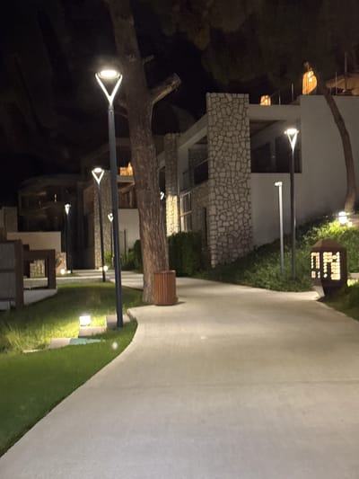 This media file captures an outdoor scene at night, likely in Bodrum, Turkey, showcasing the exterior of a modern building complex. The building features a combination of white walls and stone-clad sections, with balconies and visible windows. Several outdoor lights are strategically placed, illuminating pathways, gardens, and architectural elements.
Prominent objects include a large tree trunk in the foreground, its branches silhouetted against the night sky and the illuminated building. A series of modern, upward-facing pole lights line a paved walkway and a grassy area. One light fixture, with a distinctive triangular metal frame, is clearly visible. A wooden trash receptacle is also situated near the walkway. The lighting suggests a warm, inviting ambiance.
The scene appears to be a tranquil evening or night. There are no people visible, indicating a quiet or private setting. The presence of well-maintained landscaping, including manicured grass and strategically placed lighting, suggests a resort, hotel, or upscale residential area. One object displays text: a small, cubic sign with illuminated dots forming letters or numbers, though the text is not entirely decipherable. The overall impression is one of a serene and aesthetically pleasing outdoor space designed for evening enjoyment.