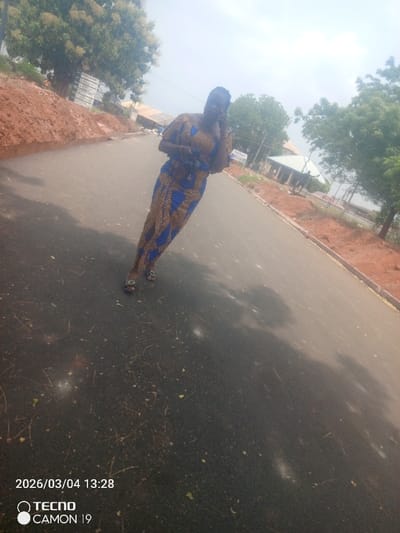 The image, dated 2026/03/04 13:28 and captured by a TECNO CAMON 19, depicts an outdoor scene in Nsugbe, Nigeria. The primary subject is an adult female standing on an asphalt road that diagonally recedes from the lower left to the upper right. She wears a full-length, patterned traditional dress featuring blue, brown, and gold geometric motifs, and dark open-toed sandals with metallic embellishments. Her hair is styled in braids. She holds a dark object in her left hand and a mobile phone to her right ear with her right hand, suggesting active communication. The road surface is dark asphalt with scattered light-colored debris. On both sides of the road are elevated verges of red-brown earth, appearing recently disturbed. In the background, numerous green trees are visible, including a large tree with light-colored blossoms on the left. Several distant buildings, one with a light-colored roof, are partially obscured by foliage. A small white sign with blue text is positioned on the right verge. The sky appears bright and hazy.