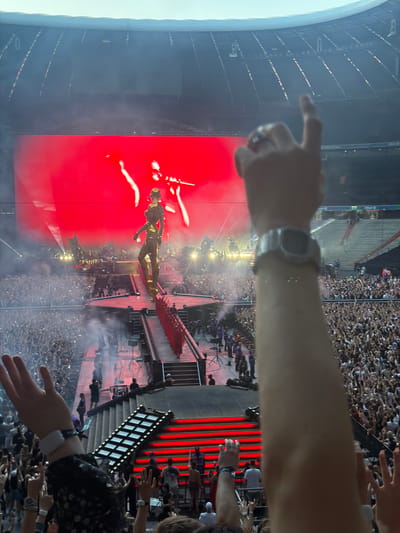 This media file captures a concert or performance event at a large stadium, likely in Munich, Germany, given the provided location context. The scene is dominated by a massive LED screen displaying a vibrant red background, with a stylized figure of a person in a dark outfit performing on stage. Smoke effects add to the dramatic atmosphere of the performance.
In the foreground, several people are visible, holding up their hands and phones, suggesting they are attendees enjoying the show. One person's arm, wearing a watch, is prominently featured, extending into the frame. Another person's hand is also visible, with fingers pointed towards the stage, possibly indicating engagement with the performance or pointing out something specific. The audience is packed into the stadium seating, with many people holding up lights from their phones, creating a sea of small lights amidst the crowd.
The event appears to be taking place in the evening or at night, as evidenced by the stage lighting and the illuminated elements within the stadium. The overall mood seems to be one of excitement and shared experience, with the collective focus directed towards the performer and the spectacle on stage. The architectural elements of the stadium, with tiered seating and prominent railings, are partially visible, providing context for the grand scale of the event.