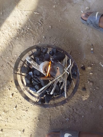 This overhead shot depicts an outdoor scene in Keffi, Nigeria, focusing on a circular metal charcoal burner placed directly on light brown, sandy ground mixed with small pebbles and debris. The burner, constructed from concentric rings of metal wire or rebar, contains numerous irregularly shaped pieces of black charcoal. A small fire is active within the burner, characterized by an orange-yellow flame emanating from a small, dark, possibly metallic or ceramic object placed amidst the charcoal and thin, light-colored wooden kindling splinters.
To the upper right of the burner, the bare foot and lower leg of an individual, wearing a dark grey sandal with a white Nike swoosh logo, are partially visible. Another leg, also wearing a similar dark grey Nike sandal, is partially visible at the bottom center-right of the frame. The ground shows subtle indentations and minor scattered detritus. Strong natural light from an unseen source casts a distinct shadow diagonally across the upper left portion of the image, indicating daylight conditions.