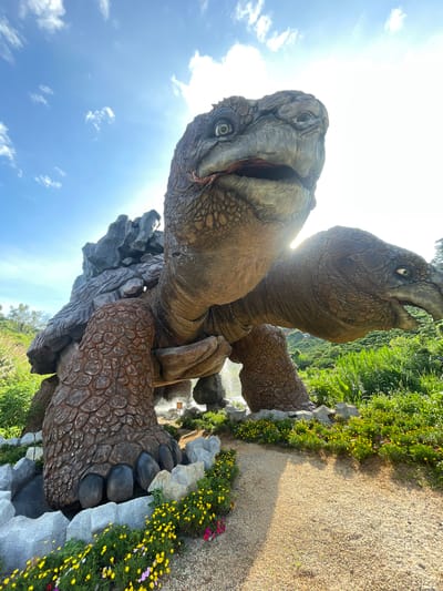 The image shows a large, realistic statue of a tortoise. The tortoise is posed with its head raised and mouth open, appearing to be looking up at the sky. Its shell is textured and appears to be made of rock-like material. The statue is situated outdoors, with a pathway leading towards it. The surrounding environment consists of lush green vegetation and a bright blue sky with scattered white clouds. The sun is visible, casting rays of light that create a lens flare effect. The statue appears to be a prominent feature in a park or tourist attraction, as indicated by the landscaping and pathway. The location is identified as Thôn 1, Vietnam, suggesting it's a specific spot within that region.