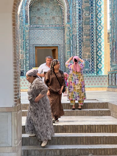 The image shows an outdoor courtyard with intricate blue and turquoise tilework on the walls of a building, likely a mausoleum, in Samarkand, Uzbekistan. In the foreground, four people are visible on a set of stone steps. On the left, a woman in a patterned grey and white dress and headscarf is looking down. Next to her, a woman in a leopard-print dress and headscarf is looking to the right. Behind them, a man in a white collared shirt is holding a smartphone and looking towards the right. Further back and to the right, a woman in a colorful floral dress and pink headscarf is posing with her hands on her head. A young girl in a pink dress is partially visible behind the woman in the floral dress. The overall atmosphere suggests a tourist visit to a historical and culturally significant site.
